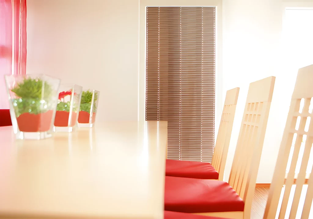 Eine Tür-Innenjalousie in einem freundlich eingerichtetem Wohnzimmer, hinter einem hellen Tisch mit Pflanzen und Sesseln